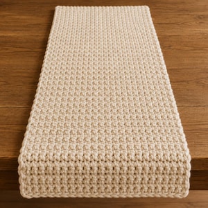 May include: A cream-colored, rectangular crochet table runner. The handmade runner is placed on a wooden table. The crochet pattern is visible, adding texture to the piece. This table runner is suitable for home decor.