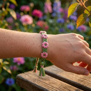 May include: A green and pink macrame bracelet featuring pink flower accents. The bracelet is worn on a wrist and has a tassel with pink and green threads. The background is a blurred view of a garden with colorful flowers.