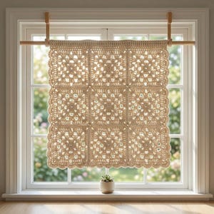 Op de afbeelding: Een vierkant beige gehaakt paneel bestaande uit negen aan elkaar verbonden granny squares. Het stuk is voorzien van een geschulpte rand en hangt aan een lichtgekleurde houten roede in een kozijn.