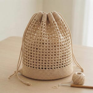 Puede incluir: Una bolsa de ganchillo beige con cordón, que presenta un patrón de malla cuadrada abierta en la parte superior y una base sólida y densa. La bolsa incluye cordones de ajuste beige a juego en ambos lados y una pequeña bola de hilo.
