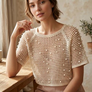 Puede incluir: Top de crochet color crema con mangas cortas y cuello redondo. La parte superior está adornada con adornos de perlas dispersas. El diseño de tejido abierto crea un aspecto delicado y aireado. La parte superior es una prenda elegante.