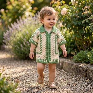 Può includere: Un bambino sorridente indossa una giacca all'uncinetto color crema e verde con colletto e maniche corte, che cammina su un sentiero di pietra. La giacca ha un'abbottonatura frontale e finiture verdi. Il bambino indossa sandali marroni.