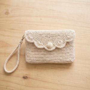 May include: A beige crocheted clutch bag. It has a scalloped edge on the flap, a button closure, and a matching wrist strap. The handmade bag is likely used for carrying small items.