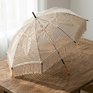 May include: A cream-coloured, openwork crochet umbrella with a wooden handle and black accents. The umbrella has a fringe trim and leaf-like patterns. It is on a rustic wooden table.