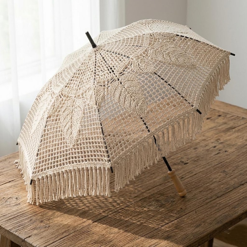 May include: A cream-coloured, openwork crochet umbrella with a wooden handle and black accents. The umbrella has a fringe trim and leaf-like patterns. It is on a rustic wooden table.