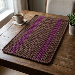 May include: A rectangular, brown crocheted placemat with two vertical purple stripes. The placemat is placed on a wooden table, with a cup of tea and a small succulent plant in the background. The texture of the crochet is visible.