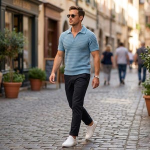 May include: A light blue short-sleeved polo shirt with a textured knit design. The shirt is paired with black pants and white sneakers. The person is wearing sunglasses and a watch. The background shows a cobblestone street and buildings.