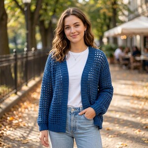 May include: A woman wearing a blue open-weave cardigan over a white t-shirt and blue jeans. The cardigan has a V-neck and long sleeves. The background shows a park with trees and a blurred cafe.
