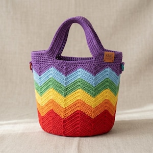May include: A handmade crochet tote bag with a rainbow chevron pattern. The bag features a purple handle and top, transitioning through blue, green, yellow, orange, and red. A small brown tag with "KANE CROCHET" is attached.