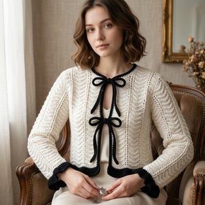May include: Cream crochet cardigan with black trim and bow fastenings. The long-sleeved sweater has a textured stitch pattern and a scalloped edge. A vintage-style garment.