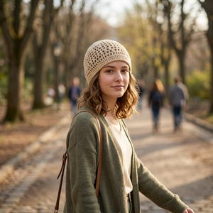 May include: A woman wearing a beige crochet beanie and a green cardigan. The beanie has an open weave design. The cardigan is a muted green color. A brown leather strap bag is over the shoulder. The background is a blurred park scene with trees and people.