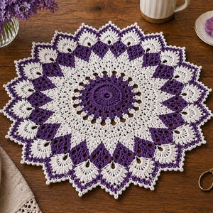 May include: A handmade, circular crochet doily in white and purple. The doily features a central purple circle, surrounded by white and purple diamond-shaped patterns. The outer edge is scalloped. This decorative item is perfect for adding a touch of handmade charm to any home.