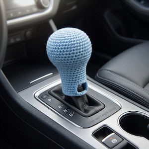 May include: A light blue crocheted gear shift knob cover. The cover is made of a soft, textured yarn and fits over the gear shift knob in a car. The cover has a loop at the bottom.