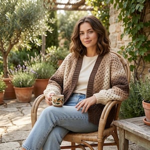 May include: A crochet cardigan in shades of brown and cream, with a V-neck design. The open-weave pattern creates a textured look. The cardigan is worn with a cream-colored top and blue jeans. A ceramic mug with a leaf design is held in the hand.
