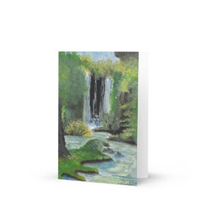 Op de afbeelding: Gevouwen wenskaart met een aquarel schilderij van een waterval. Het kunstwerk gebruikt tinten groen, blauw en wit om het gebladerte en het water weer te geven. De kaart is van binnen leeg. De schildering toont een waterval.
