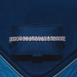 May include: A tennis bracelet featuring a continuous row of clear, pear-cut stones set in a metallic band. The bracelet is displayed horizontally, showcasing the uniform arrangement of the faceted, teardrop-shaped stones that reflect light.
