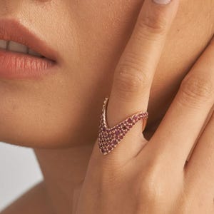 May include: A rose gold ring with a V-shaped design, set with small, round, red gemstones. The ring is on a person's finger, with the hand and part of the face visible.