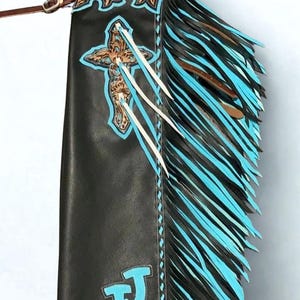 Op de afbeelding: Een zwarte leren chap met lange turquoise en zwarte franjes aan de zijkant. Het item is versierd met een bruin, bloemrijk bewerkt lederen kruis met een turquoise omlijning, een zilveren ster-concho aan de bovenkant en turquoise letters JJ onderaan.