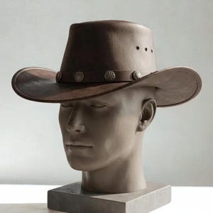May include: A brown leather cowboy hat featuring a wide brim and a decorative band adorned with circular silver-toned metal studs. The hat includes three small ventilation eyelets on each side of the crown, providing a structured and classic look.