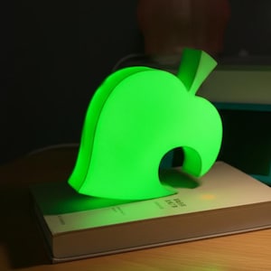 Op de afbeelding: Een heldergroen blad-vormig nachtlampje en een witte bureaulamp staan op een houten oppervlak. Het bladlicht gloeit, en de bureaulamp heeft een ronde kop. Een boek en een turquoise spelconsole staan op de achtergrond.