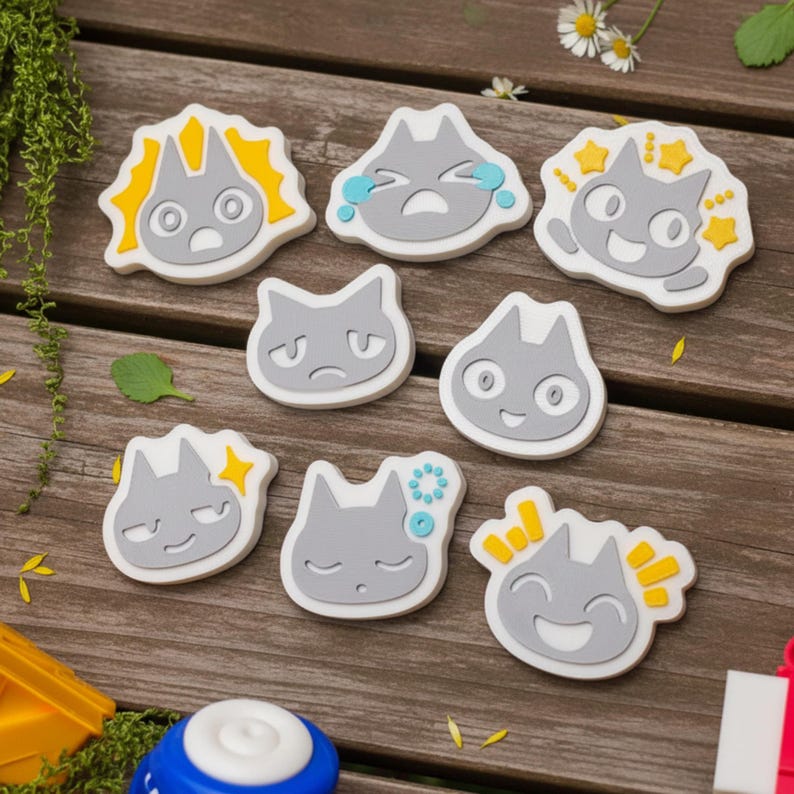 May include: A collection of gray cat-shaped stickers with various facial expressions, including happy, sad, and surprised. Each sticker has a white border and yellow or blue accents. The stickers are arranged on a wooden surface.