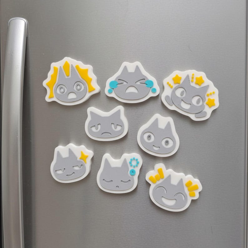 May include: A collection of gray cat-shaped magnets with various facial expressions, including crying, smiling, and surprised. Each magnet has a white border and yellow or blue accents. The magnets are attached to a silver refrigerator door.