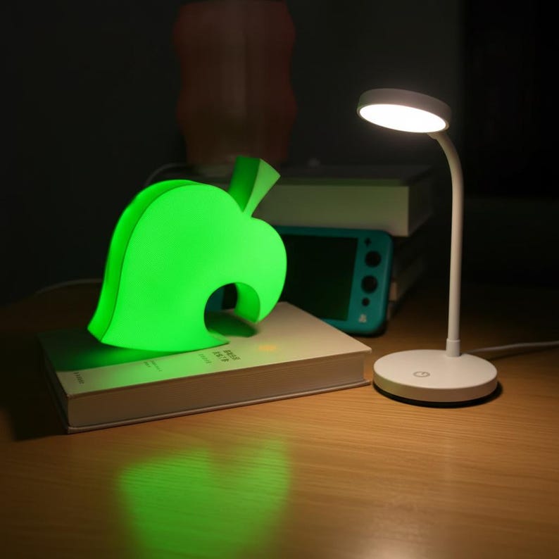 Op de afbeelding: Een heldergroen blad-vormig nachtlampje en een witte bureaulamp staan op een houten oppervlak. Het bladlicht gloeit, en de bureaulamp heeft een ronde kop. Een boek en een turquoise spelconsole staan op de achtergrond.