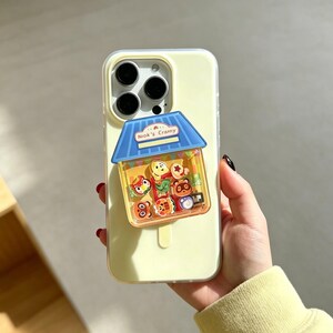 May include: A yellow phone case with a blue and yellow house-shaped charm. The charm features cartoon characters and the text "Niok's Cranny". The phone case is held by a person.
