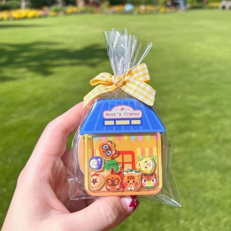 May include: A clear cellophane bag tied with a yellow gingham bow, containing a colorful, house-shaped trinket box. The box features the text "Nook's Cranny" and contains various character-shaped candies. The background is a blurred green lawn.