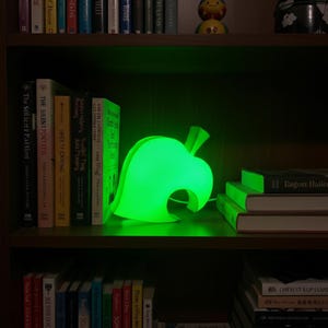 Op de afbeelding: Een heldergroene, bladvormige lamp, die doet denken aan een videogame-icoon, staat op een houten plank. De lamp straalt een helder groen licht uit en cre&euml;ert een zachte gloed. De plank maakt deel uit van een donkere houten boekenkast.