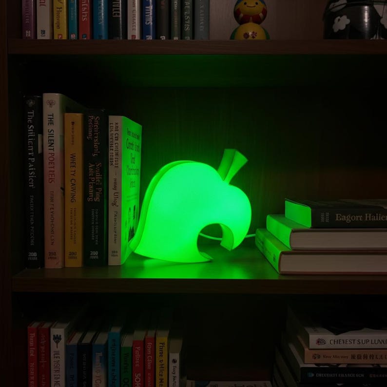Op de afbeelding: Een heldergroene, bladvormige lamp, die doet denken aan een videogame-icoon, staat op een houten plank. De lamp straalt een helder groen licht uit en cre&euml;ert een zachte gloed. De plank maakt deel uit van een donkere houten boekenkast.