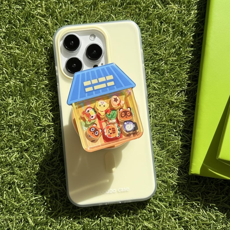May include: A light yellow phone case with a blue and yellow house-shaped phone grip. The grip is filled with colorful cartoon character charms. The phone case is on a green surface.