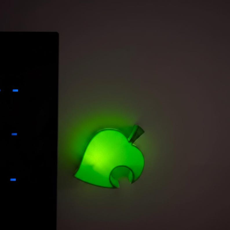 Op de afbeelding: Een gloeiende groene blad-vormige lamp. Het licht is aan een muur bevestigd en straalt een heldere, levendige gloed uit. Het ontwerp is een gestileerd blad, waarschijnlijk voor decoratieve doeleinden. De achtergrond is neutraal van kleur.