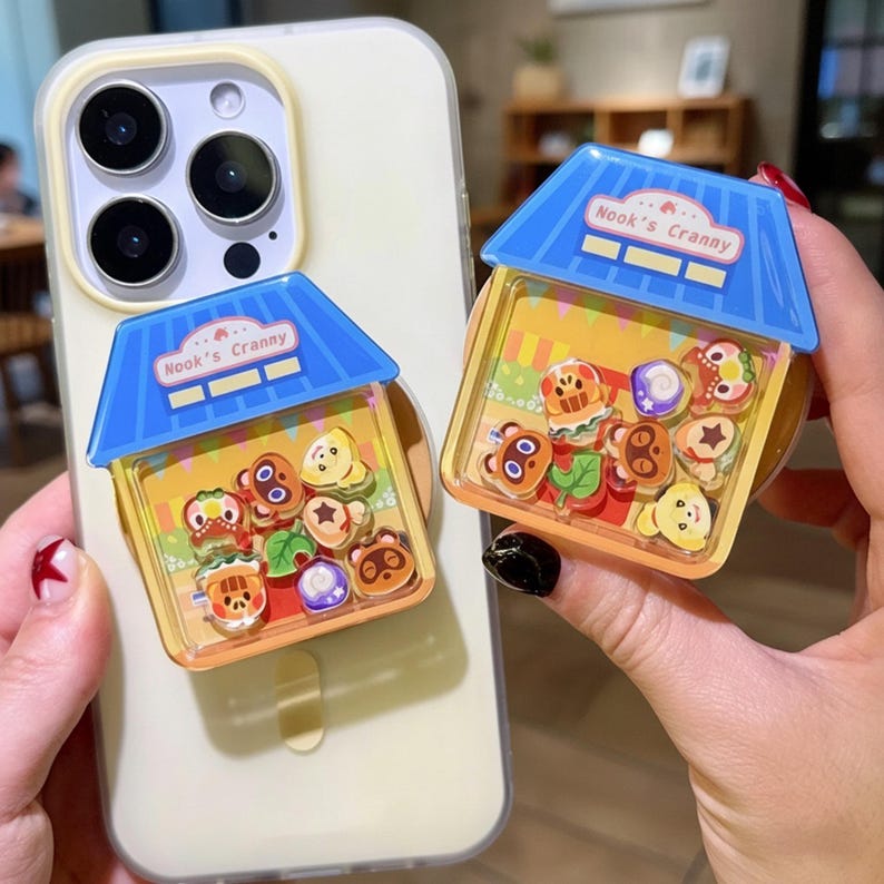 May include: Two phone grips featuring a miniature house design with the text "Nook's Cranny" on a blue roof. The clear plastic houses contain colorful character charms. One grip is attached to a phone.