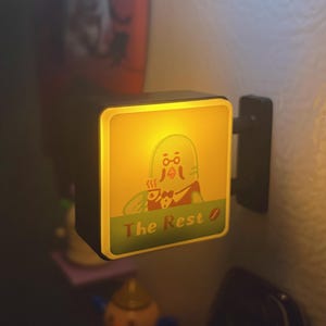 May include: A black, square-shaped illuminated sign with a cartoon figure holding a coffee cup. The sign reads "The Rest" in green text, with a yellow glow emanating from within. The sign is mounted on a wall.