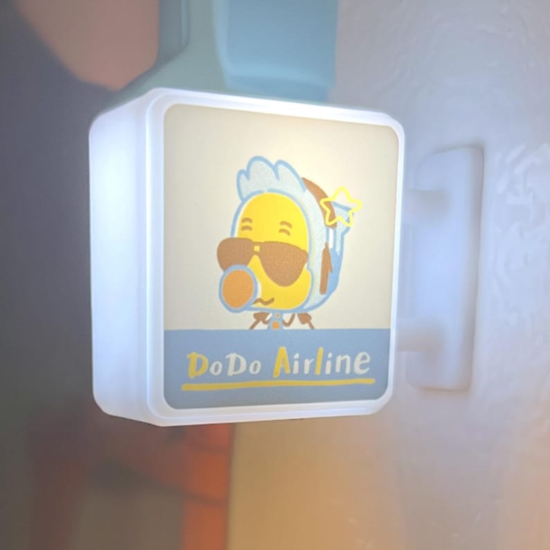 May include: A square, illuminated sign featuring a cartoon character wearing sunglasses and a bonnet. The sign has the text "DoDo Airline" in a blue and yellow color scheme. The sign is mounted on a white wall.