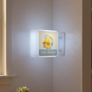 May include: A square, illuminated wall light fixture with a cartoon image of a yellow bird wearing sunglasses and a blue and white hat. The text "Do Do Airline" is below the image. The light is mounted on a white wall.
