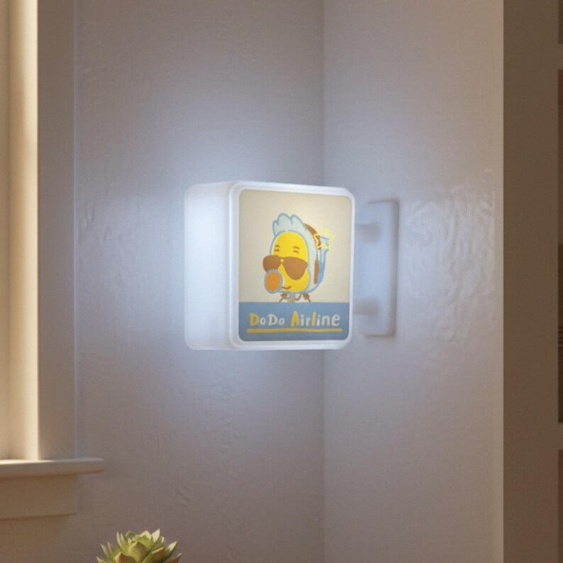 May include: A square, illuminated wall light fixture with a cartoon image of a yellow bird wearing sunglasses and a blue and white hat. The text "Do Do Airline" is below the image. The light is mounted on a white wall.