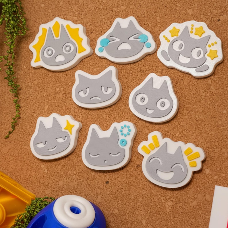 May include: A collection of gray cat-shaped stickers with various facial expressions. Each sticker has a white border and yellow or blue accents. The stickers are arranged on a brown surface, with a few green sprigs visible.
