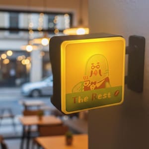 May include: Illuminated sign with a cartoon figure holding a cup, the text "The Rest" is displayed on a green banner. The sign is square with a black frame and is mounted on a wall. The sign is lit with a warm yellow glow.