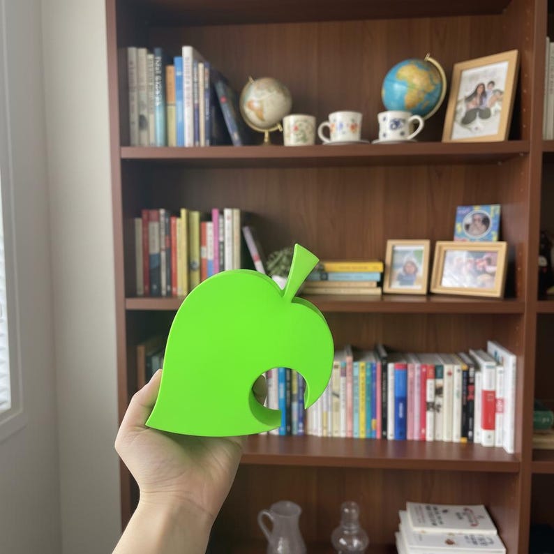 Op de afbeelding: Een heldergroen, blad-vormig object, ge&iuml;nspireerd op de Animal Crossing-game, wordt voor een boekenplank gehouden. Het object heeft een glad oppervlak en een uitgesneden ontwerp. De achtergrond toont een houten boekenkast met boeken, globes en ingelijste foto's.