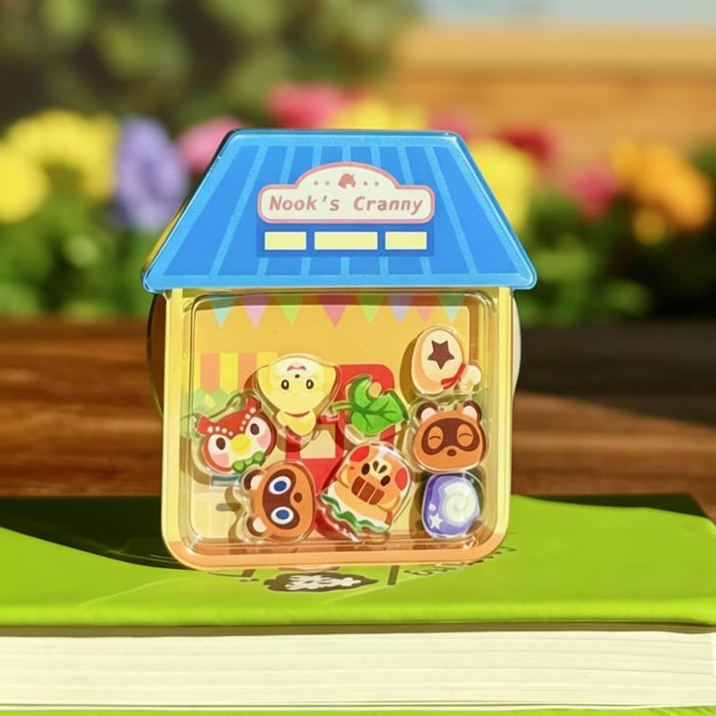 May include: A clear plastic house-shaped container with a blue roof and the text "Nook's Cranny". Inside are several character charms from the Animal Crossing video game series. The container sits on a green book.