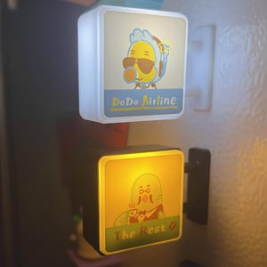 May include: Two illuminated square signs with cartoon graphics. The top sign reads "DoDo Airline" and features a yellow bird wearing sunglasses. The bottom sign says "The Rest" and shows a cartoon character with a cup. Both signs have a white border.
