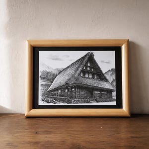 Könnte beinhalten: Eine gerahmte Schwarz-Weiß-Zeichnung eines traditionellen japanischen Bauernhauses mit einem steil abfallenden Strohdach. Das Kunstwerk ist in einem hellen Holzrahmen gefasst und ruht auf einer Holzoberfläche.