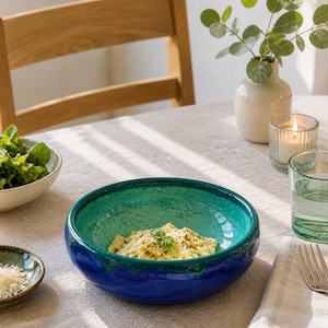 May include: A round ceramic bowl featuring a deep blue exterior and a speckled turquoise interior. The bowl contains a serving of pasta topped with a small green herb leaf. The glaze on the rim shows a distinct drip pattern where the colours meet.