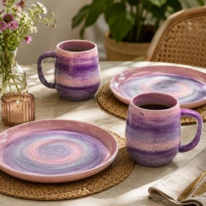 Peut inclure: Deux tasses en céramique et deux assiettes assorties présentant un motif aquarelle tourbillonnant dans des tons de violet, rose et bleu. La vaisselle a une texture mouchetée et une finition brillante, disposée sur des sets de table tressés.
