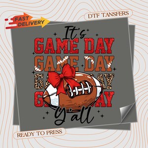 May include: A graphic design featuring a football with a red bow, the words "It's GAME DAY y'all" in red glitter and brown lettering. The design is ready to press.