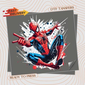 May include: A DTF transfer sheet featuring a graphic illustration of a red and blue superhero in a dynamic pose with a black spider emblem on the chest. The design includes white paint splatter effects and red and blue dots on a gray background.