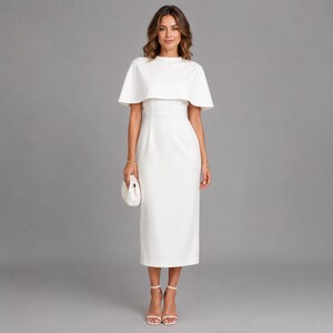 May include: A solid white midi-length dress featuring a high neckline and a structured cape-style overlay that drapes over the shoulders. The garment has a fitted bodice, a straight skirt silhouette, and is paired with a small white clutch bag.