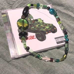 Puede incluir: Un collar de cuentas verdes, negras y transparentes, sobre una caja de CD. La portada del CD presenta una ilustración de un jeep camuflado con la palabra "Gorillaz" y una advertencia parental.